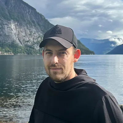 Portrait of Bence Székely, photographer, standing by a Norwegian fjord with mountains in the background