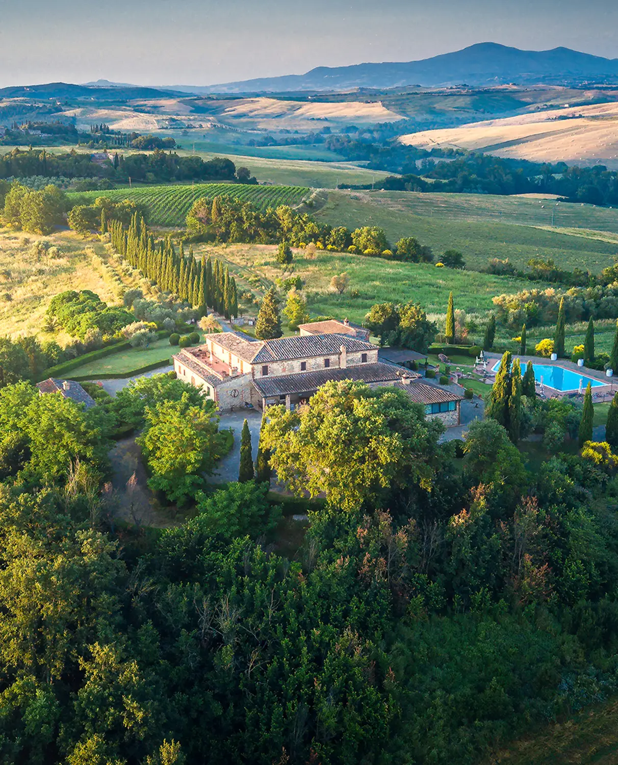 Aerial view of a hotel in Tuscany surrounded by vineyards and cypress trees at golden hour. Drone photography portfolio image.